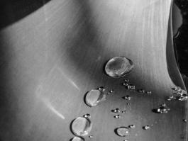 Close-up of water droplets on a smooth, dark surface, creating a serene and reflective mood. Light softly highlights the droplets' curvature. 